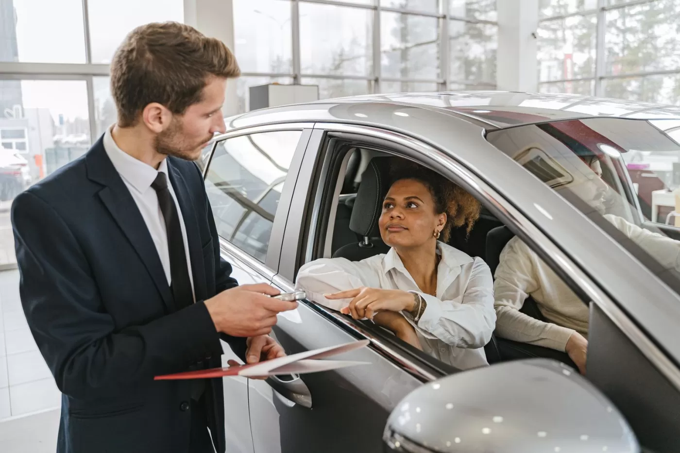 A salesperson handing paperwork to a smiling customer seated in a new car